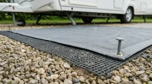 Close-up photorealistic photograph of a multi-layer groundsheet setup on a UK seasonal caravan pitch hardstanding, showing a black breathable underlay mesh beneath a durable grey main groundsheet and gravel.