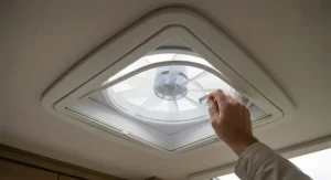 Macro detail of a hand detaching the fine-mesh flyscreen from a caravan roof vent fan, designed to keep midges and insects out of the living area.