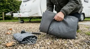 A high-resolution 4K photorealistic photograph of a neatly folded heavy-duty grey-charcoal durable groundsheet being slipped into a sturdy, zipped polyethylene storage bag on a gravel pitch.