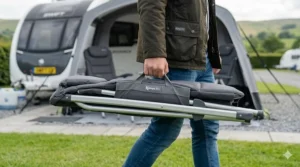 A person carrying a compact folded caravan chair by its integrated handle across a grassy campsite.