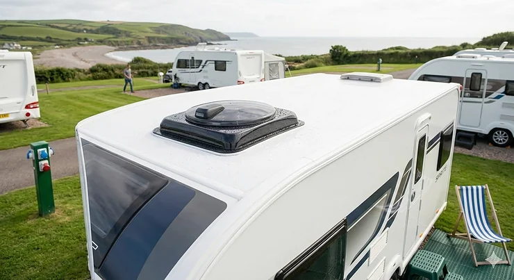 High-angle professional photograph of a modern white touring caravan featuring an installed roof vent fan with an integrated rain sensor, parked at a scenic UK coastal campsite. roof vent fan with rain sensor for caravans