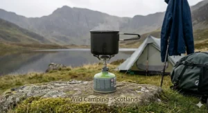 A photorealistic wide-angle illustration of a lightweight gas stove in use during a misty Scottish Highland wild camping trip on a lochan shore.