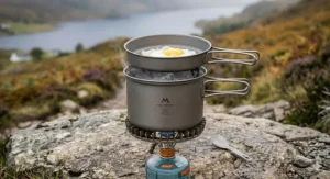 A photorealistic illustration showing a hard-anodised aluminium cookware set for backpacking utilising an integrated metal heat-exchange ring at its base to capture the stove flame for increased fuel efficiency.