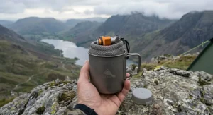 A detailed close-up showing a human hand holding a packed ultralight titanium backpacking stove and pot kit on a rocky ridge in the Lake District, showcasing its compact size.
