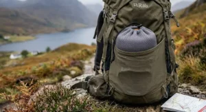 A photorealistic illustration showing an entire 9-piece cookware set for backpacking meticulously nested inside its grey mesh bag and tucked into the side pocket of a technical hiking rucksack.