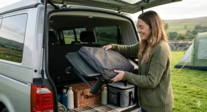 A compact, lightweight camping grill being easily packed into the boot of a silver campervan for a weekend trip in the British countryside.