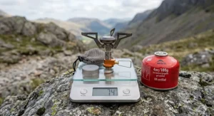 A titanium ultralight backpacking stove resting on a precise digital scale displaying 74 grams, positioned on a rugged rock with a Lake District valley background.