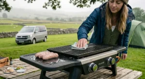 A durable, stainless steel camping grill being used during typical drizzly British weather, with a camper wiping down the surface in the Lake District.