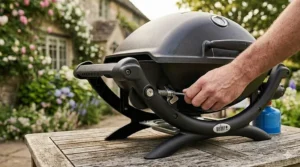 A detailed photorealistic 4K close-up showing a hand interacting with the spring-loaded locking pin and lever of the folding stand mechanism of a Weber portable BBQ on a weathered wooden table.