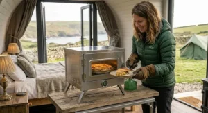 Alt text for image 7: A photorealistic 4K photograph capturing the interior of a luxury glamping pod in Cornwall. The woman from the hero image, wearing her green Rab jacket and using brown leather gloves, carefully places a baked fruit cobbler, baked inside the stainless steel camping oven, onto a ceramic plate. The glamping pod's comfortable interior and distant coastal views of rolling hills and dry stone walls are visible through the doors, with a green bell tent softly blurred in the distance. Small, detailed wisps of steam are rising from the hot cobbler.