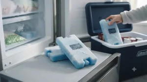 Two heavily frosted, frozen blue ice blocks being transferred from a domestic freezer shelf into the open cool box, highlighting the buildup of ice crystals.