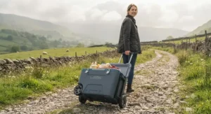 A person in a wax jacket effortlessly pulling the grey wheeled cool box along an uneven, pebbled country path.