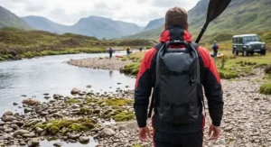 Paddler wearing a black dry bag backpack with padded shoulder straps while walking along a gravel riverbank.
