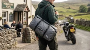 Rider carrying a waterproof motorcycle dry bag using a padded shoulder strap near a cafe.